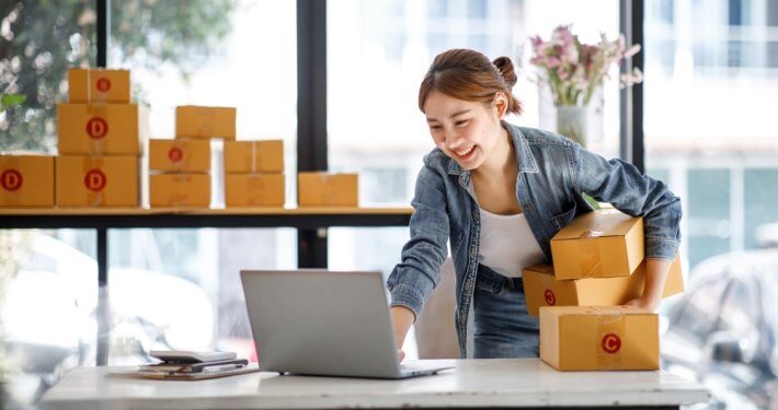 business owner smiling while holding boxes and looking at laptop