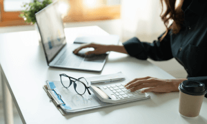 woman sitting at a desk and typing on a laptop with her right hand and on a calculator with her left hand